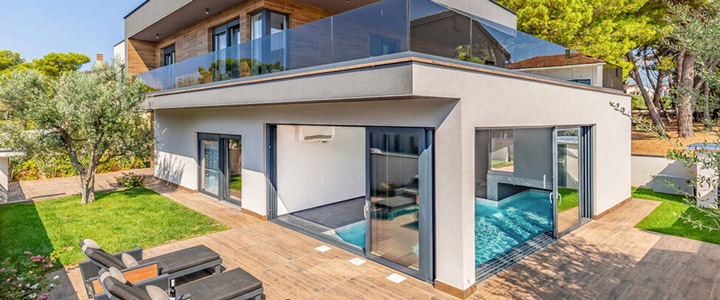Ein stilvolles, modernes Ferienhaus mit viel Glas und warmen Holzflächen liegt inmitten grüner Natur. Im Inneren öffnet sich ein ruhiger Innenpool im lichtdurchfluteten Raum.